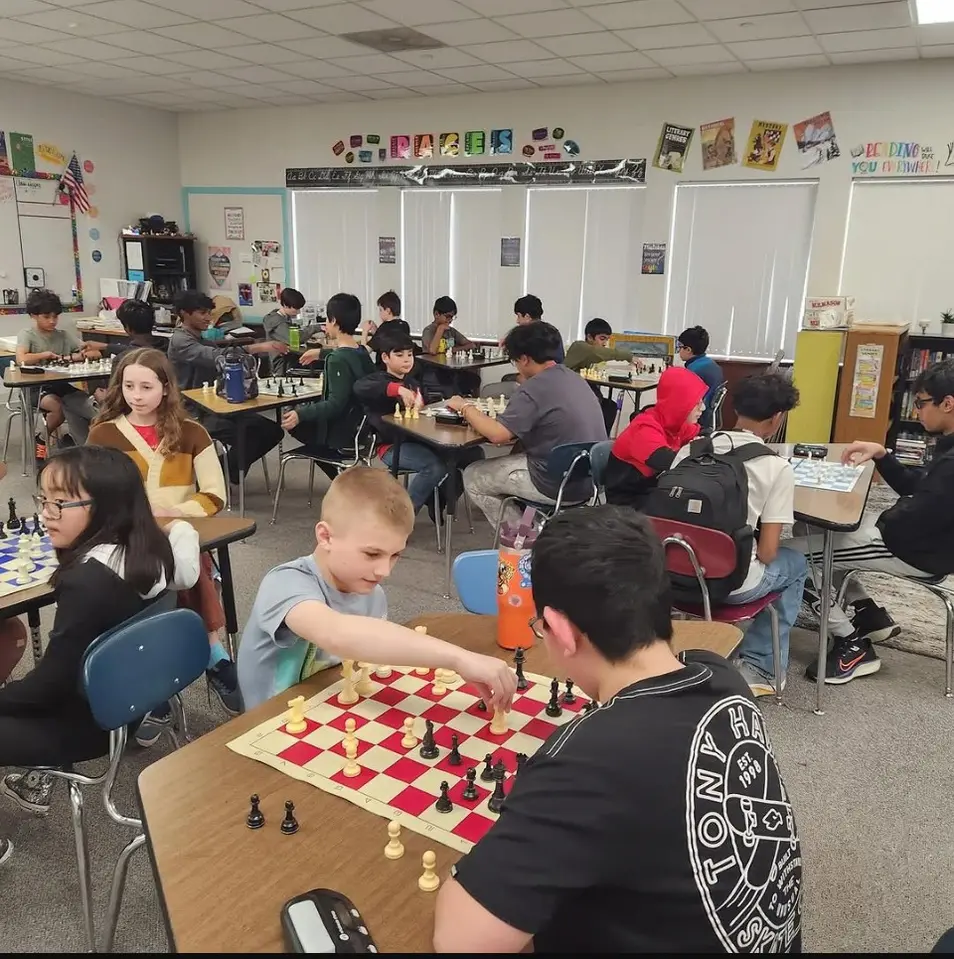 A classroom of students at desks, one reaching across a red-and-white vinyl chessboard in the foreground.